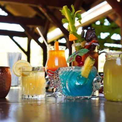 Assorted colorful cocktails with fruit garnishes on a wooden bar in an outdoor setting.