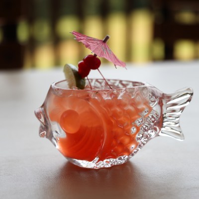 Fish-shaped glass with a pink drink, lime slice, cherries, and umbrella on a table.