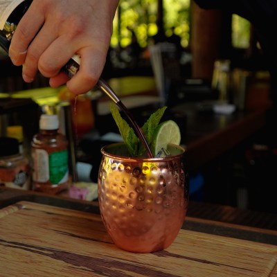 Hand pouring drink into copper mug with mint and lime on wooden board.